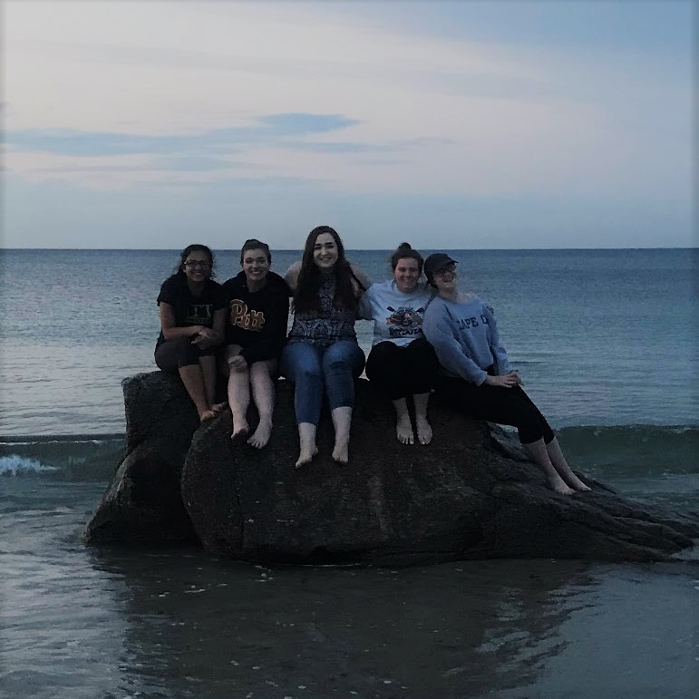 A group of five teenagers sitting on a large rock on the shore.